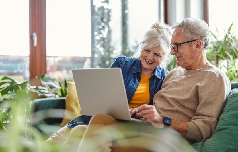 A smiling senior couple relaxing on a sofa in their home while using their laptop computer.